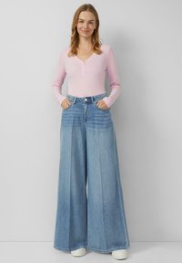 Jasnoróżowa bluzka henley z długim rękawem i trzema guzikami, zestawiona z szerokimi jasnoniebieskimi dżinsami i białymi trampkami.