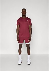 Mies, joka seisoo maroonin värisessä Puma-urheilupaidassa ja -shortseissa, valkoisissa sukissa ja valkoisissa lenkkareissa, yksinkertaista valkoista taustaa vasten.