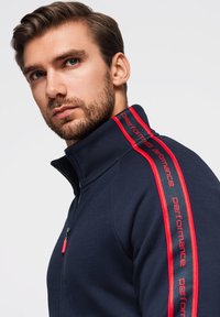 Marinefarbener Zip-Up-Jacke mit roten "Performance"-Streifen auf den Schultern. Weicher Stoff, hoher Kragen und ein roter Reißverschluss-Zug. 