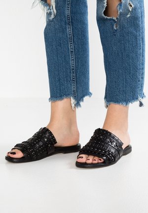 Pieds portant des sandales slip-on en cuir noir clouté et un jean en denim bleu à ourlet effiloché avec des déchirures au-dessus des genoux sur un sol blanc.