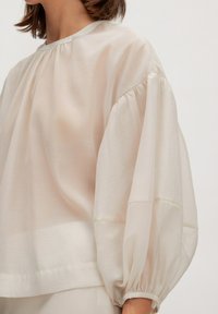 Helle beige Bluse mit lockerer Passform, gerafftem Rundhalsausschnitt und voluminösen, puffärmeligen Ärmeln. Hergestellt aus einem weichen, durchsichtigen Stoff.