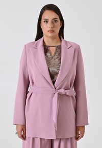 Lichtroze blazer met lange mouwen, geknoopte revers en een strik in de taille. Gekleed over een gestructureerde bruine top met een keyhole-halslijn.