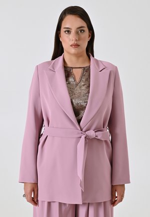 Blazer rosa chiaro con maniche lunghe, rever a punta e vita legata. Indossato sopra una maglia marrone testurizzata con scollatura a chiave.