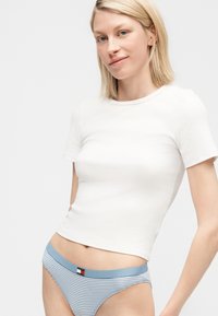 Ξανθιά γυναίκα που φοράει εφαρμοστό λευκό κοντομάνικο μπλουζάκι και μπλε-λευκά ριγέ εσώρουχα με μπλε ζώνη μέσης.