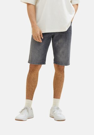 Homme portant un short en jean noir délavé, des baskets blanches, des chaussettes blanches et une chemise blanche ample, debout, une main dans la poche.