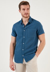 Camisa de manga corta abotonada en azul profundo con un tejido texturizado. Cuenta con un bolsillo en el pecho y sutiles rayas verticales.