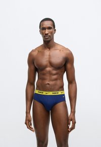 Μπλε ναυτικό ανδρικό εσώρουχο Nike με κίτρινη ζώνη μέσης που φέρει το λογότυπο, εφαρμοστή σχεδίαση και απαλή υφή υφάσματος. Εμφανίζεται σε μοντέλο.