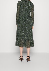 Mujer vistiendo un vestido midi negro con un pequeño estampado floral azul y mangas largas transparentes, combinado con sandalias de tacón negro, vista trasera.