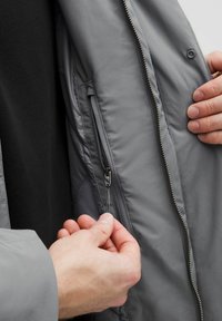 Käed tõmbavad halli jaka sisetasku tõmblukk sisse, mille all on must särk.