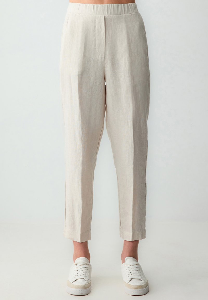 Pantaloni in lino beige chiaro con una vestibilità rilassata, caratterizzati da una cintura elastica, tasche laterali e orli leggermente svasati. Abbinati a sneaker bianche.