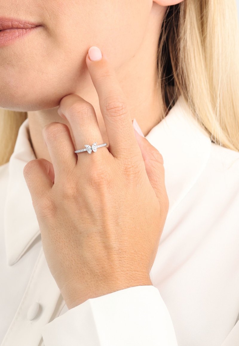 Anello d'argento con pietra ovale scintillante e intricati dettagli laterali, indossato su un dito vicino a un volto, caratterizzato da pelle liscia e unghie di colore chiaro.