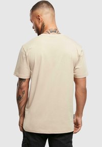 Beige t-shirt met korte mouwen en een ronde hals, gladde textuur en een label aan de zoom. Heeft zichtbare tatoeages op de arm.
