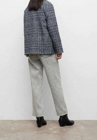 Person in einer blauen karierten Jacke, hellgrauen Jeans und schwarzen Stiefeletten steht vor einer schlichten weißen Wand.