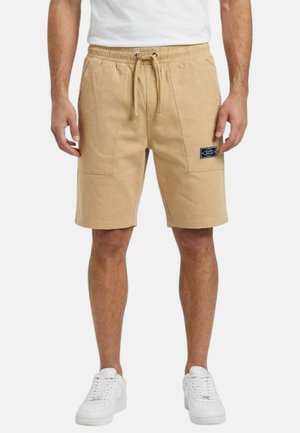 Homme portant un short beige à cordon avec poches frontales et des baskets blanches, debout devant un fond uni.