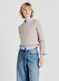 Beige gestrickter Pullover mit Rundhalsausschnitt, über einem hellblauen Hemd getragen. Dazu werden lockere, hoch sitzende Jeans aus Denim getragen. Einfarbiger Hintergrund.