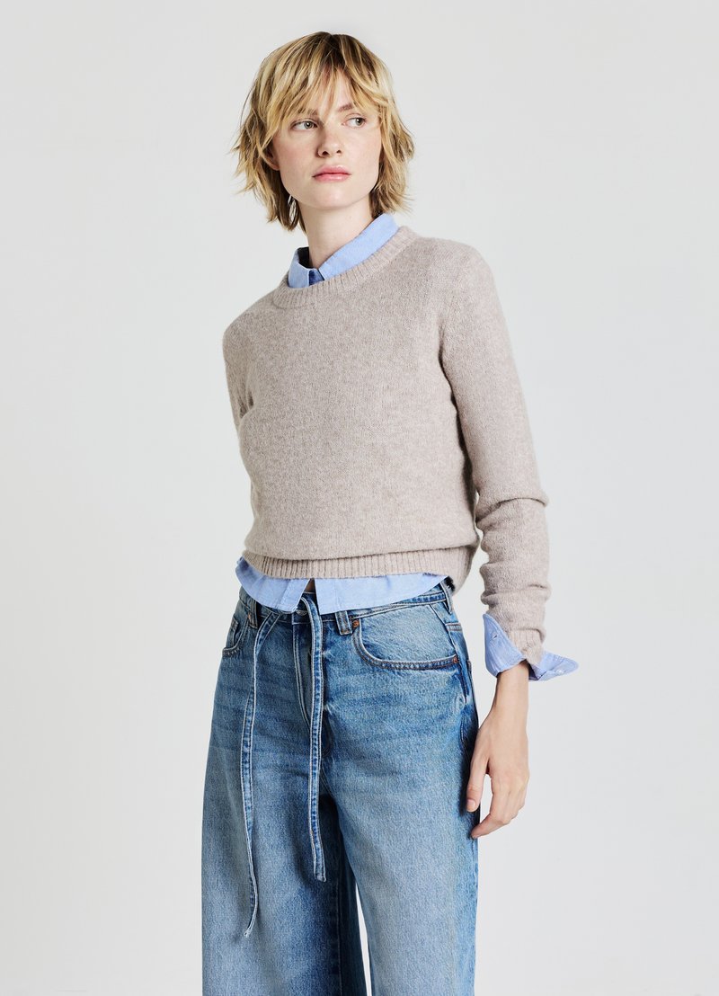 Pull en maille beige à col rond, superposé à une chemise bleu clair. Porté avec un jean en denim ample taille haute. Fond uni.