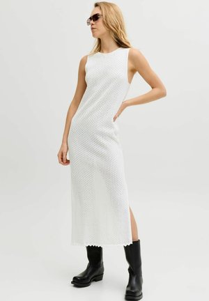 Kobieta w białej, bezrękawnikowej, teksturowanej sukience midi, czarnych botkach i ciemnych okularach przeciwsłonecznych, pozująca z ręką na biodrze.