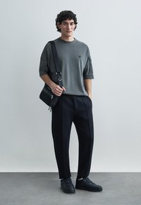 Šedá oversized košile s krátkými rukávy, černé plisované kalhoty a černé tenisky. Malé logo na košili; crossbody taška se zipovým detailem.