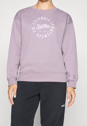 Osoba nosící světle fialovou mikinu Columbia Sportswear a černé kalhoty s viditelnými logy Columbia.