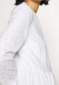 Weißes Kleid aus strukturiertem Baumwollstoff mit einem gestickten Muster. Verfügt über lange Ärmel und einen ausgestellten Saum, der zarte Lochstickerei-Details zeigt.