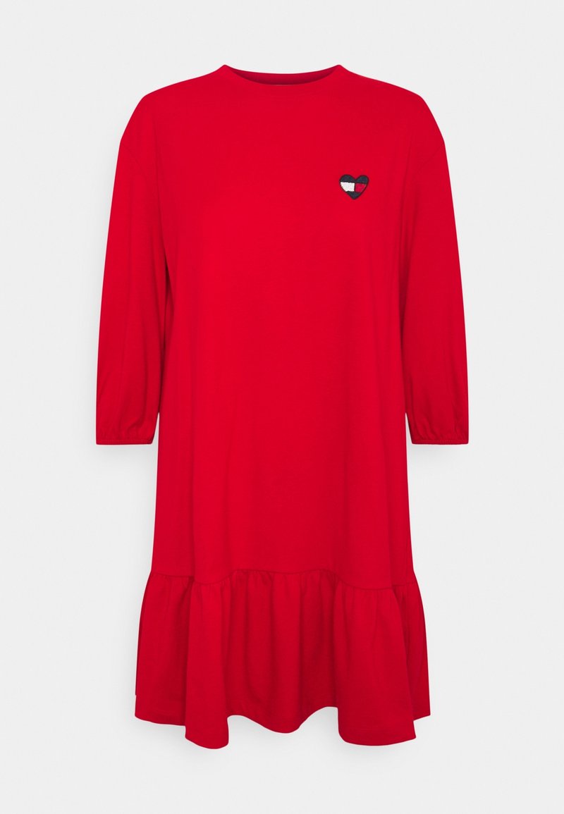 Robe rouge à manches longues avec encolure ronde, ourlet à volants et petit patch en forme de cœur bleu marine, blanc et rouge sur la poitrine gauche.