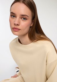 Felpa beige a girocollo con una texture morbida e spalle scese. Il tessuto liscio contrasta con i capelli lunghi e lisci della modella.