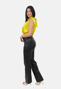 Kobieta z długimi ciemnymi włosami, ubrana w bezrękawnik z żółtego satynu, czarne skórzane spodnie i białe sandały na wysokim obcasie, pozująca z boku.