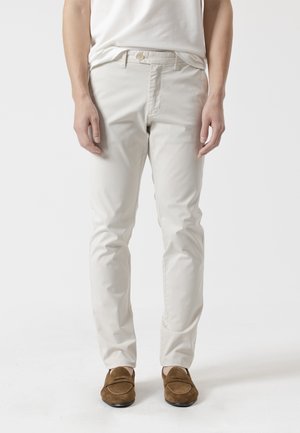 Hombre vistiendo pantalones ajustados color marfil, camiseta blanca y mocasines de ante marrón, de pie contra un fondo liso y claro.