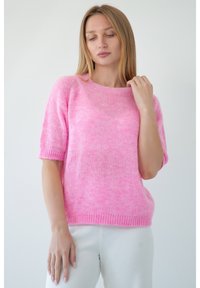 Krótki rękaw, luźny sweter z teksturowanej jasnoróżowej przędzy z prążkowanym dekoltem i brzegiem. Model nosi go z białymi prążkowanymi spodniami.