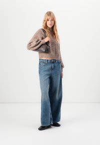 Skrojony sweter w warkoczowy splot w kolorze taupe, zestawiony z szerokimi niebieskimi dżinsami i czarnymi balerinami. Modelka trzyma szary worek, w minimalistycznym tle.