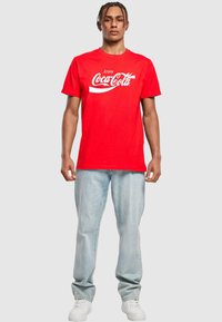 Červené bavlněné tričko s nápisem "Užívejte si Coca-Colu" v bílém písmu, doplněné o světle modré džíny a bílé tenisky.