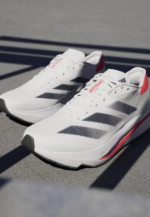 Weiße Sportschuhe mit grauen Streifen, strukturiertem Mesh-Obermaterial, roten Akzenten an der Ferse und schlankem Sohlen-Design. Schwarze Gummisohle.