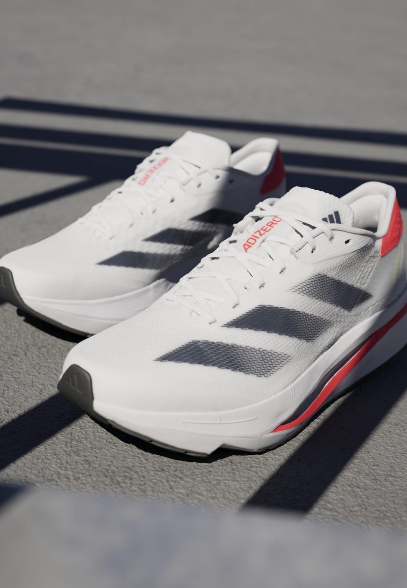 Baskets de sport blanches avec des rayures grises, tige en maille texturée, accents rouges sur le talon et design de semelle épuré. Semelle extérieure en caoutchouc noir.