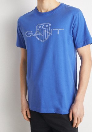Homme portant un t-shirt bleu vif à manches courtes avec un logo GANT blanc en contour et un écusson avec trois étoiles sur la poitrine.
