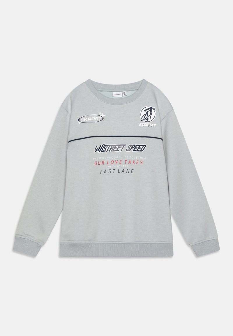 Sudadera gris clara con cuello redondo, que presenta varios logotipos e inscripciones impresas en colores contrastantes, incluyendo acentos en azul marino y rojo.