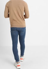 Béžový pletený svetr nosený přes modré džínové kalhoty. Oblečení má přiléhavý střih a bílé tenisky s červenými akcenty.