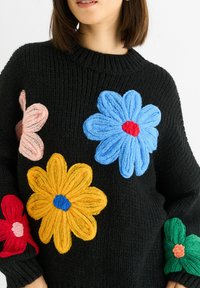 Black knitted sweater featuring three-dimensional flower appliqués in blue, yellow, pink, and green, with ribbed cuffs and neckline.