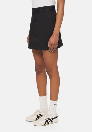 Personne portant une mini-jupe noire, des chaussettes blanches et des baskets blanches avec des rayures noires, se tenant devant un fond uni.