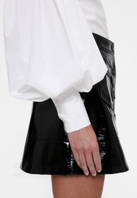 Črna mini krilo iz lakiranega usnja z bleščečo površino in razširjenim robom, združeno z belo srajco z napihnjenimi rokavi, ki je zataknjena v krilo.