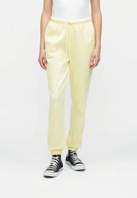 Dzelteni sporta bikses ar stringu jostu, ar apkaklēm potītēs un mīkstu tekstūru, apvienotas ar melniem augstiem sporta apaviem.