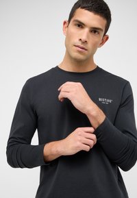 Juoda ilgomis rankovėmis medvilninė palaidinė su apvalia iškirpte ir mažu baltu "MUSTANG" logotipu ant krūtinės.