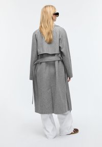 Grijze wol trenchcoat met een double-breasted ontwerp, ceintuur in de taille en een gekruiste overlap aan de achterkant, gecombineerd met witte broek en bruine schoenen.