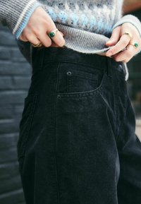 Pantaloni in corduroy neri con tasche laterali, dotati di righe verticali, vita alta e dettagli con bottoni. Indossati con un maglione leggero a fantasia.