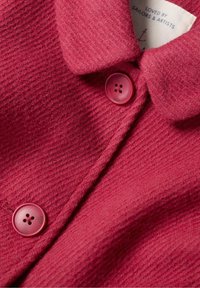 Tejido texturizado rojo con un cuello de solapa, que presenta dos botones redondos rosas. La etiqueta es visible con el texto "AMADO POR MARINEROS Y ARTISTAS."