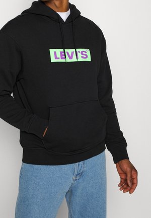 Homme portant un sweat à capuche Levi's noir avec un logo vert néon et violet et un jean bleu clair, main dans la poche du sweat à capuche, sur fond blanc.