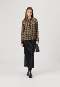 Camicia a maniche lunghe con bottoni nera con motivo geometrico dorato, abbinata a una gonna nera in satin e stivali neri con tacco, mentre tiene una piccola borsa nera.