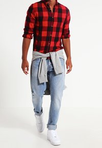 Camisa de franela a cuadros rojos y negros con botones, jeans de mezclilla azul claro y zapatillas blancas. Sudadera gris atada a la cintura.