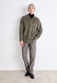 Hombre con un gorro beige, camisa de pana verde sobre un suéter negro de cuello alto, pantalones cargo grises y zapatos negros, de pie frente a una pared en blanco.
