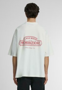 Camiseta blanca de gran tamaño con un gran estampado gráfico rojo en la parte posterior que dice "CASA MODA 1019 PROIBIZIONE" y detalles decorativos.
