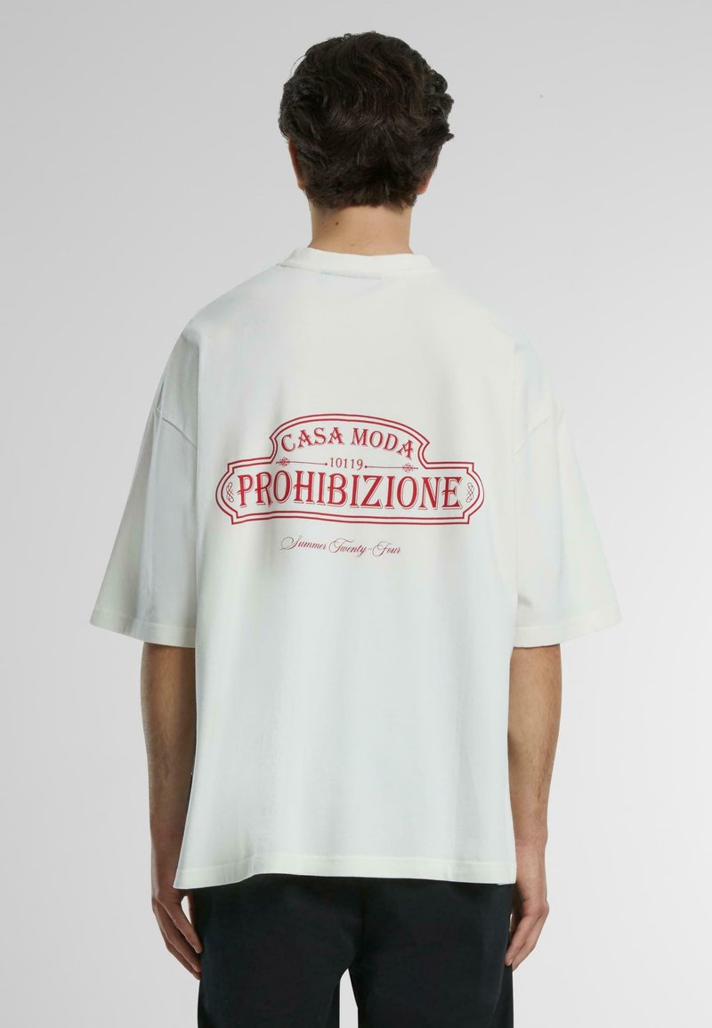 Camiseta blanca de gran tamaño con un gran estampado gráfico rojo en la parte posterior que dice "CASA MODA 1019 PROIBIZIONE" y detalles decorativos.